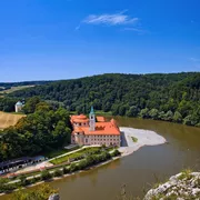 Kloster am Fluss inmitten einer grünen Landschaft unter blauem Himmel.
