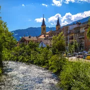 Fluss fließt durch malerische Stadt mit Kirchtürmen, Bäumen und Bergen im Hintergrund.