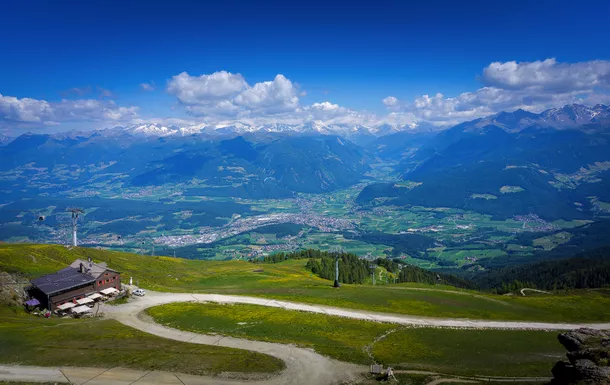 Aussicht auf alpine Landschaft mit Bergwiese, Gebäude, Serpentinenweg und bewölktem Himmel.