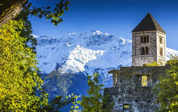 Kirchturm einer alten Steinkirche vor einer verschneiten Bergkette, umgeben von grüner Vegetation.