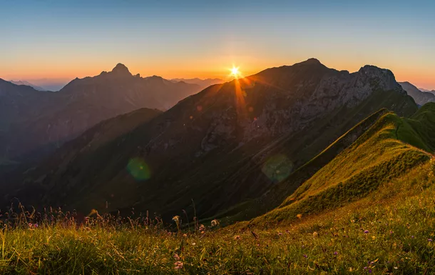 Sonnenaufgang über Bergen mit grüner Wiese im Vordergrund.