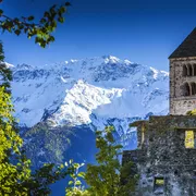 Kirchturm einer alten Steinkirche vor einer verschneiten Bergkette, umgeben von grüner Vegetation.