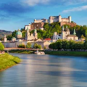 Festung Hohensalzburg über der Stadt Salzburg mit Fluss im Vordergrund bei Sonnenuntergang.