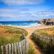 Pfad führt durch Dünenlandschaft zu einem Strand mit Felsen und blauem Himmel.