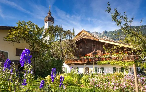 Alpenhaus mit Garten und Kirchturm im Hintergrund an einem sonnigen Tag.