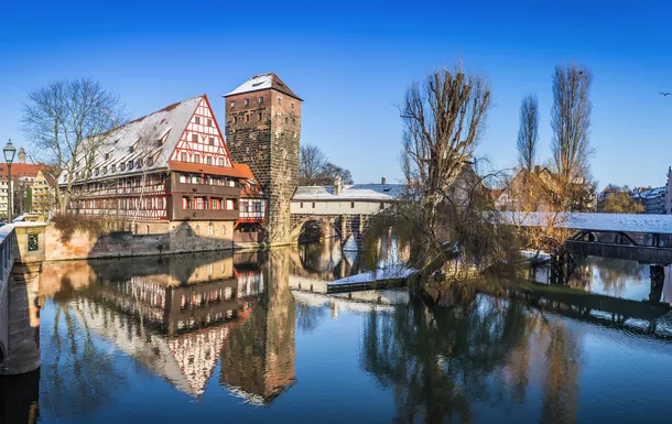 Fachwerkhaus und Brücke an verschneitem Flussufer, klarer Himmel, spiegelndes Wasser.